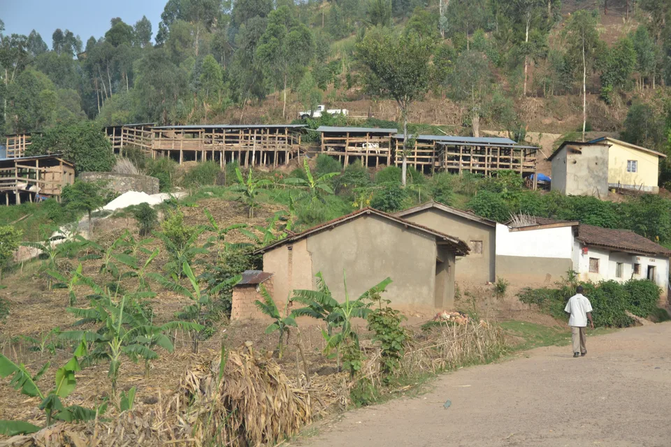 A dirt road passes hillside homes and cultivated fields, with a person walking away in the foreground.