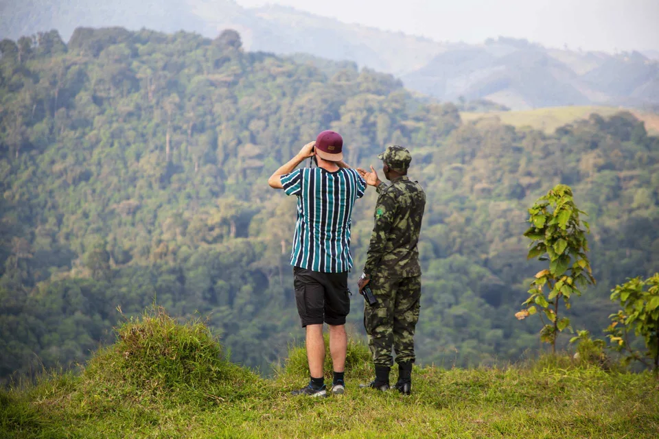 Two people looking across forested hills at Gishwati, one using binoculars.