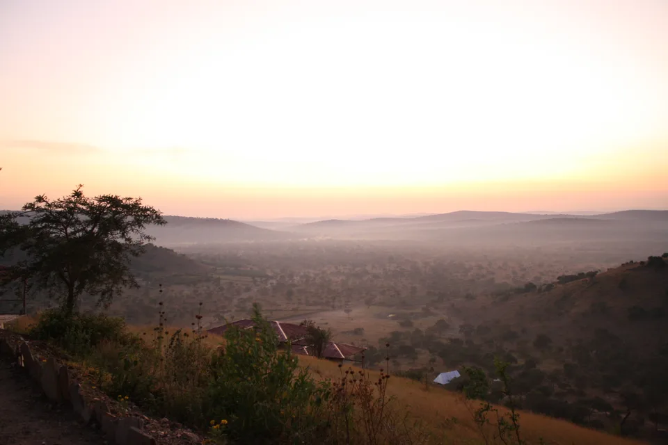 A hazy morning view across rolling woodland and grassland from a hillside near Lake Mburo, with a few roofs in the foreground.