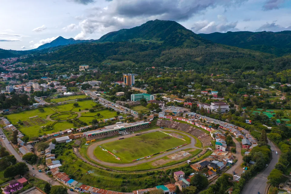 Elevated view across Mbeya’s stadium, streets and buildings toward green hills beneath cloud.