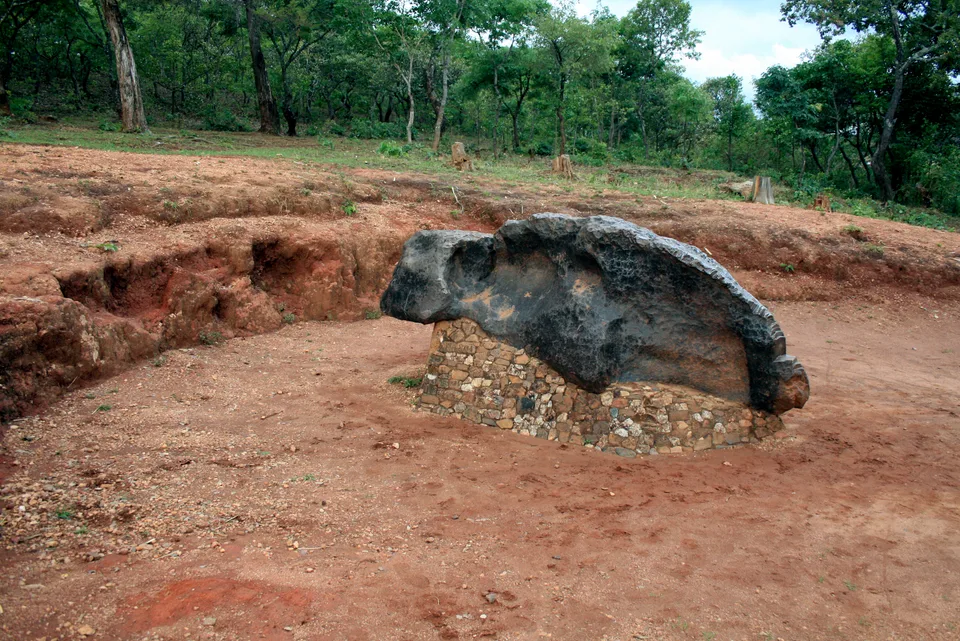 Large dark Mbozi meteorite resting on masonry support in a cleared hollow surrounded by trees.