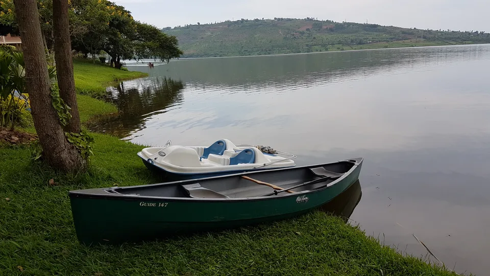 Canoe and pedal boat beside a grassy Lake Muhazi shore, with cultivated hills across the water.
