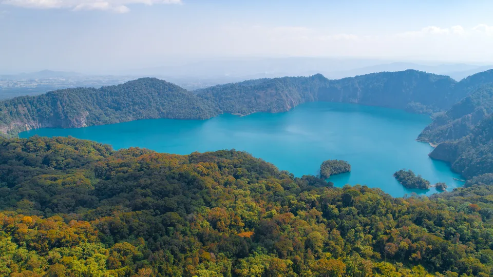Blue-green Lake Ngozi lies inside a steep forested crater rim, with small wooded islands near the shore.
