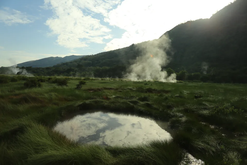 Steam rises above a green wetland with a pool and wooded hills behind it.