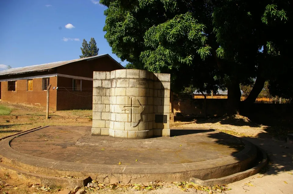 A stone memorial on a circular paved platform beneath a large tree at Ujiji, with a low building behind it.