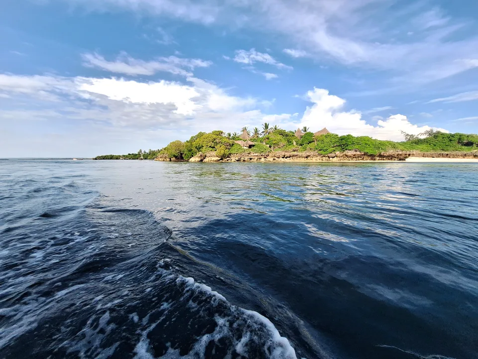 Blue water beside a low rocky shoreline, trees and thatched roofs in the Watamu area.